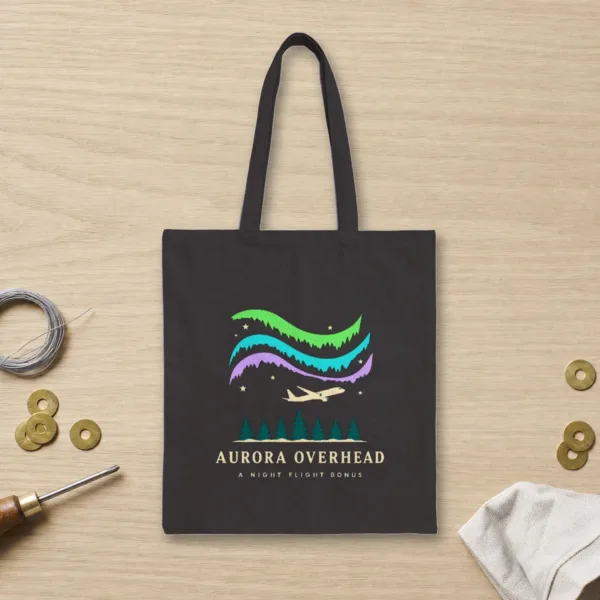 Aurora Overhead design in warm cream ink on a black canvas tote, laid flat on an oak hangar workbench