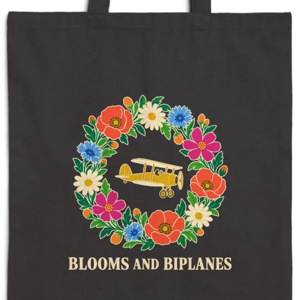 Close-up of the Blooms and Biplanes design printed in warm cream ink on a black canvas tote, with the top seam and handles