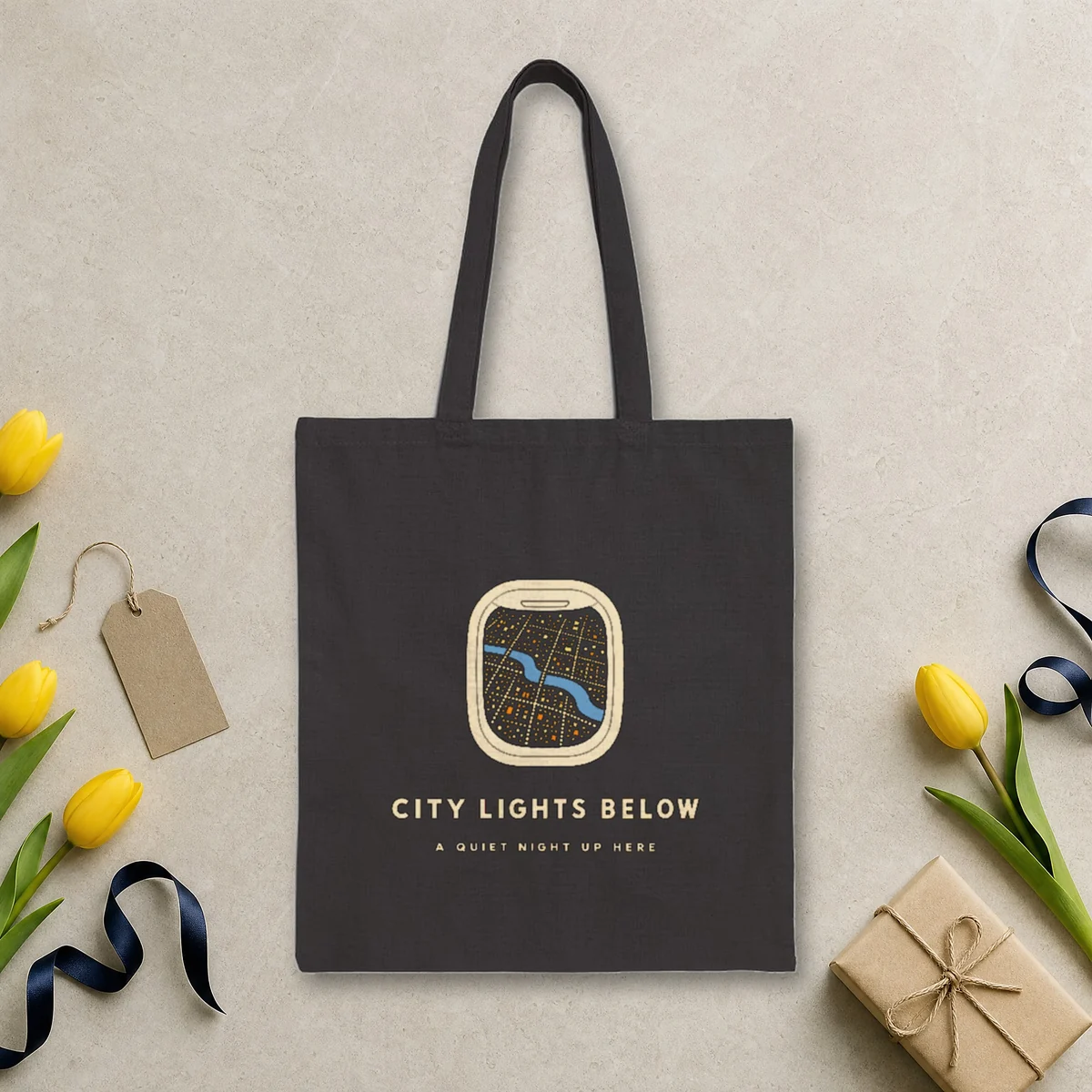 City Lights Below design in warm cream ink on a black canvas tote, laid flat on a limestone arrivals hall floor