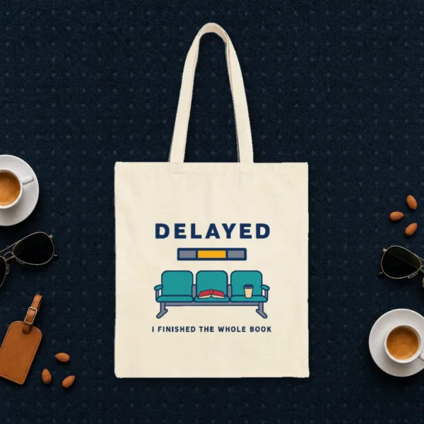 delayed-with-a-book-tote-natural-front Delayed with a Book design in navy ink on a natural canvas tote, laid flat on an airport lounge carpet