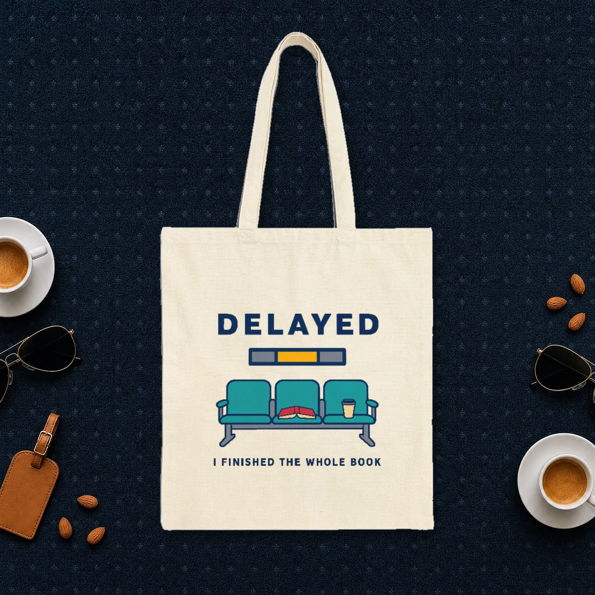 Delayed with a Book design in navy ink on a natural canvas tote, laid flat on an airport lounge carpet