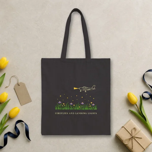 Fireflies and Landing Lights design in warm cream ink on a black canvas tote, laid flat on a limestone arrivals hall floor