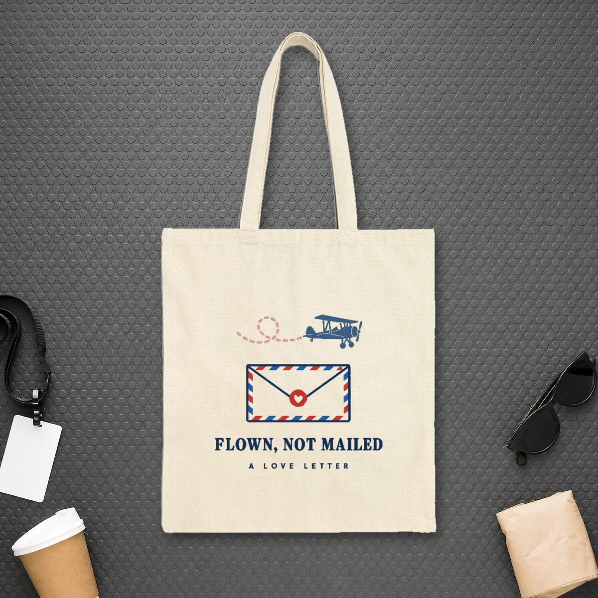 Flown Not Mailed design in navy ink on a natural canvas tote, laid flat on the studded rubber floor of a jet bridge