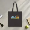 Fly-In Farmers Market design in warm cream ink on a black canvas tote, laid flat on a sunlit terminal floor by the window