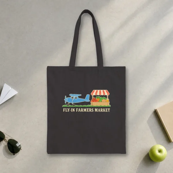 Fly-In Farmers Market design in warm cream ink on a black canvas tote, laid flat on a sunlit terminal floor by the window