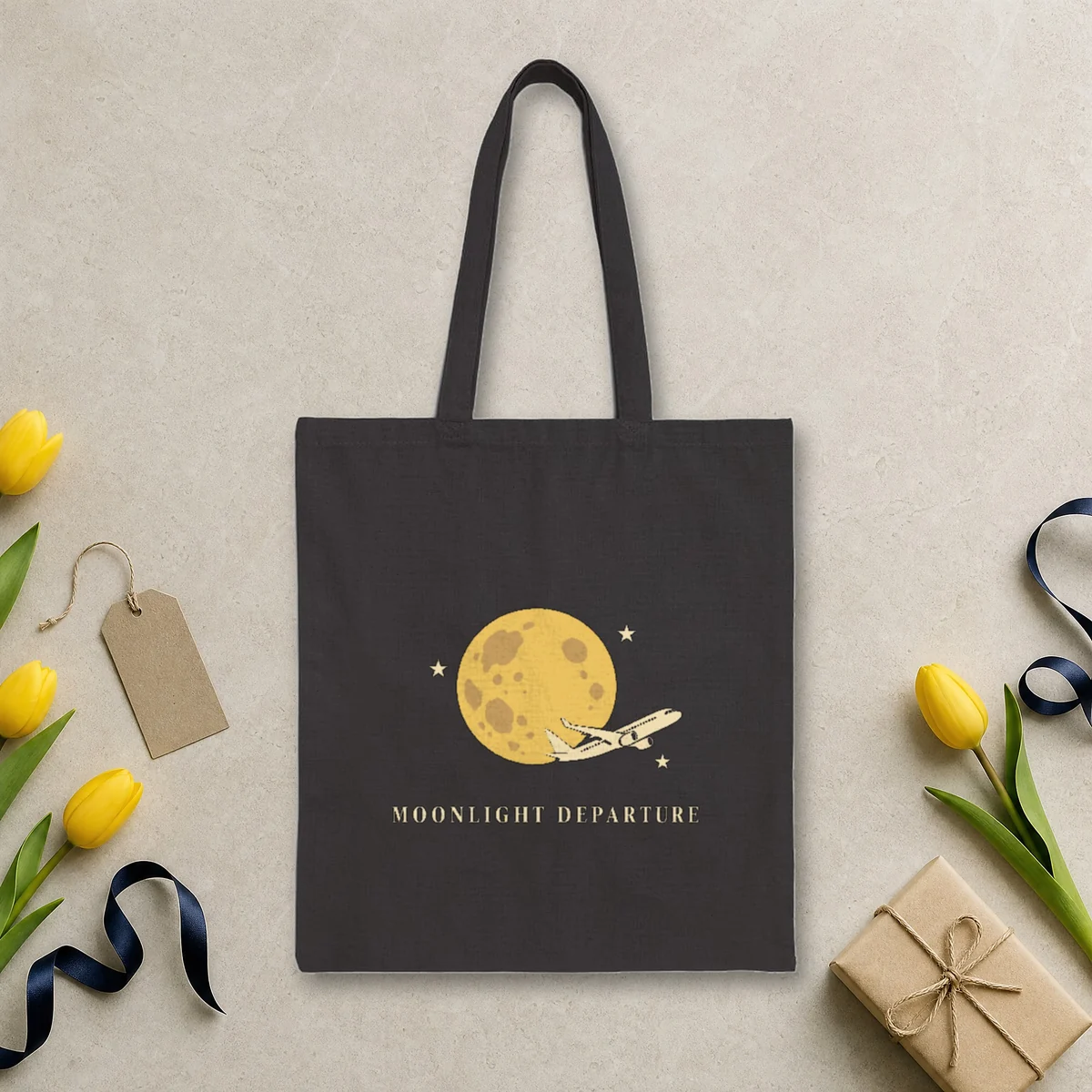 Moonlight Departure design in warm cream ink on a black canvas tote, laid flat on a limestone arrivals hall floor