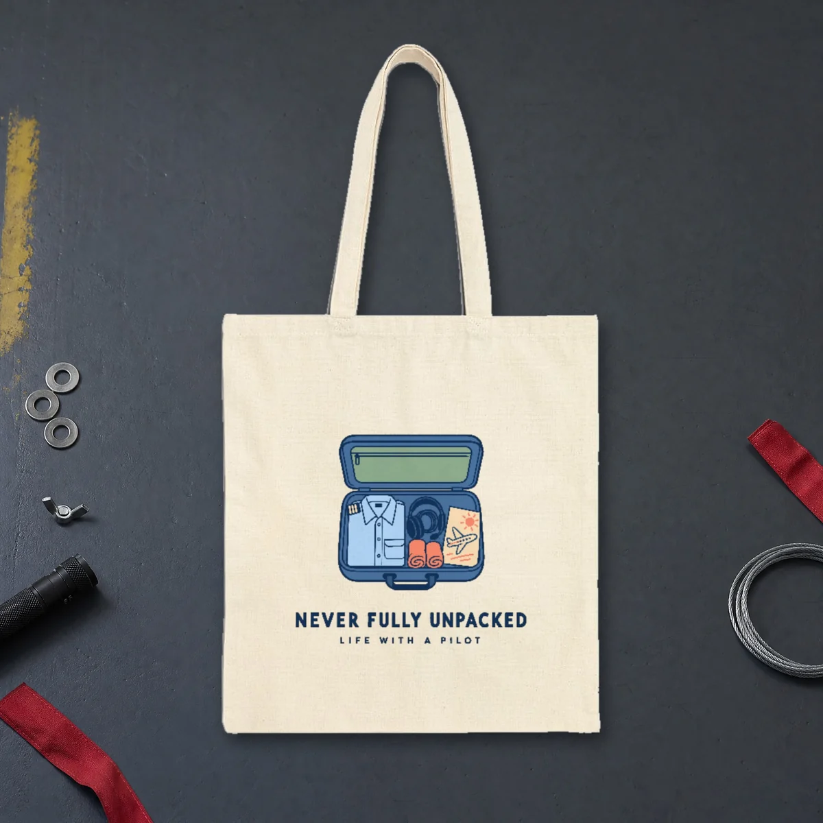 Never Fully Unpacked design in navy ink on a natural canvas tote, laid flat on a hangar floor