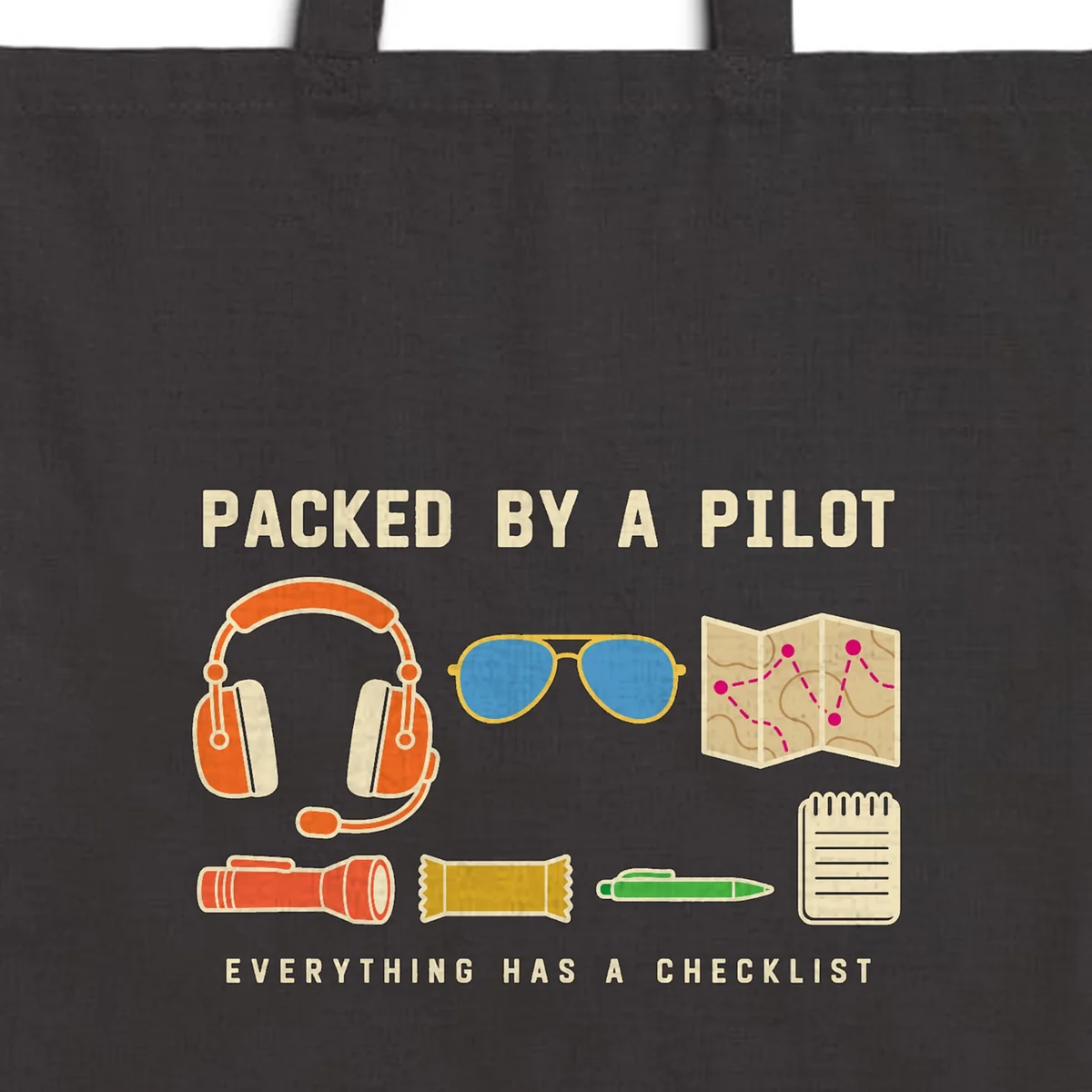 Close-up of the Packed by a Pilot design printed in warm cream ink on a black canvas tote, with the top seam and handles