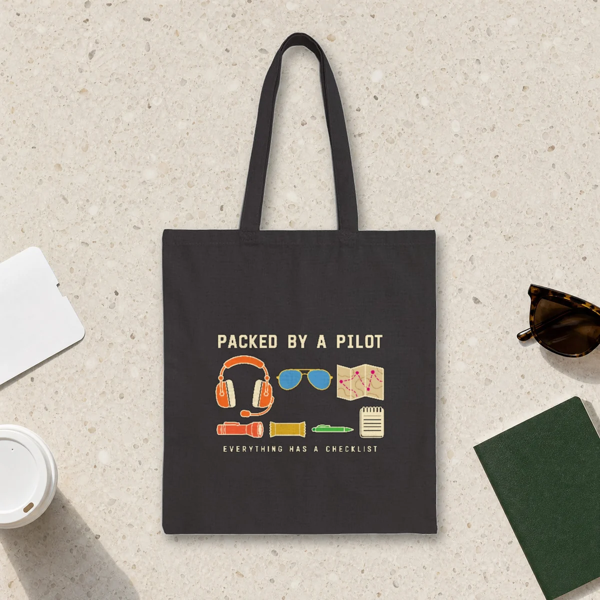 Packed by a Pilot design in warm cream ink on a black canvas tote, laid flat on a light terrazzo terminal floor