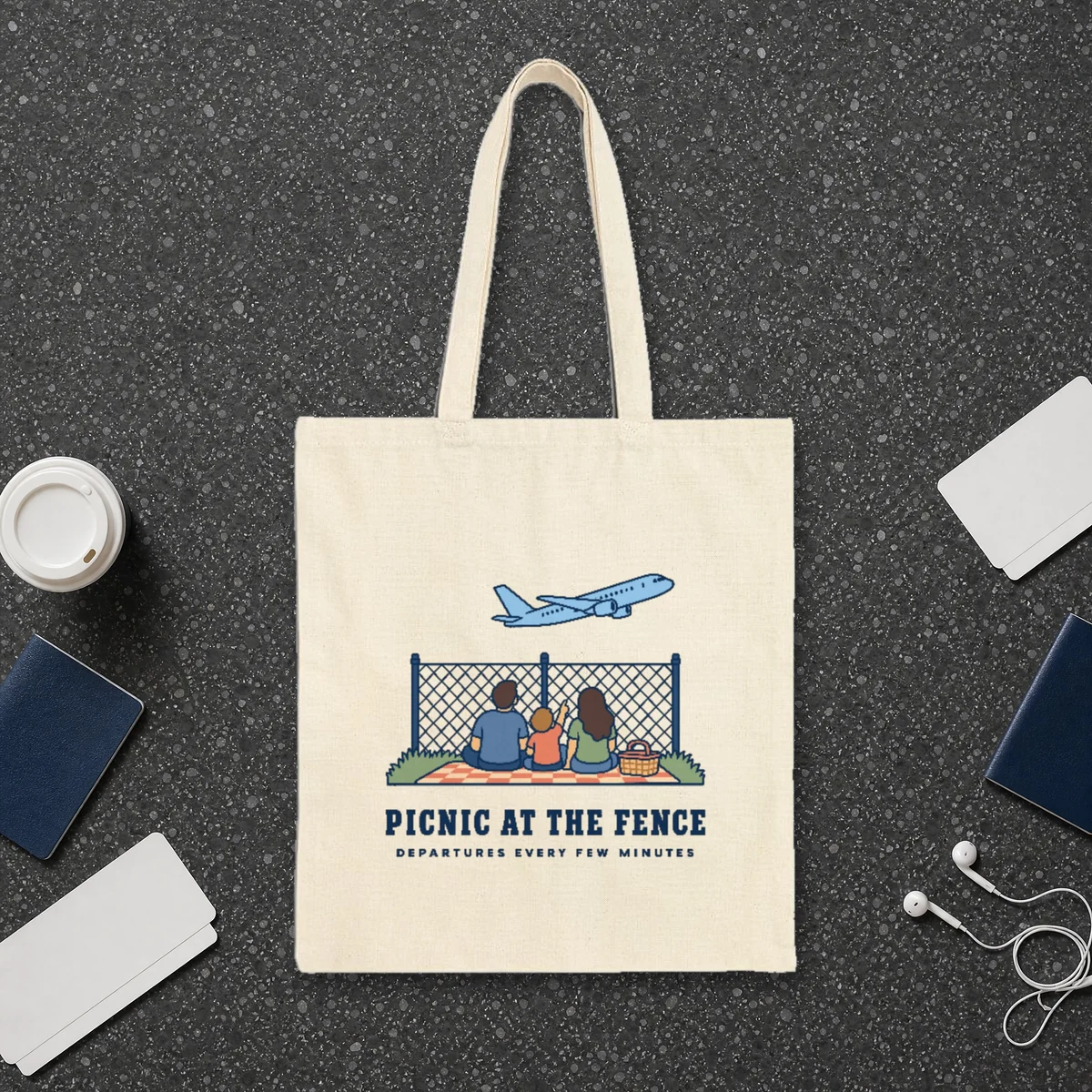Picnic at the Fence design in navy ink on a natural canvas tote, laid flat on a dark terrazzo terminal floor