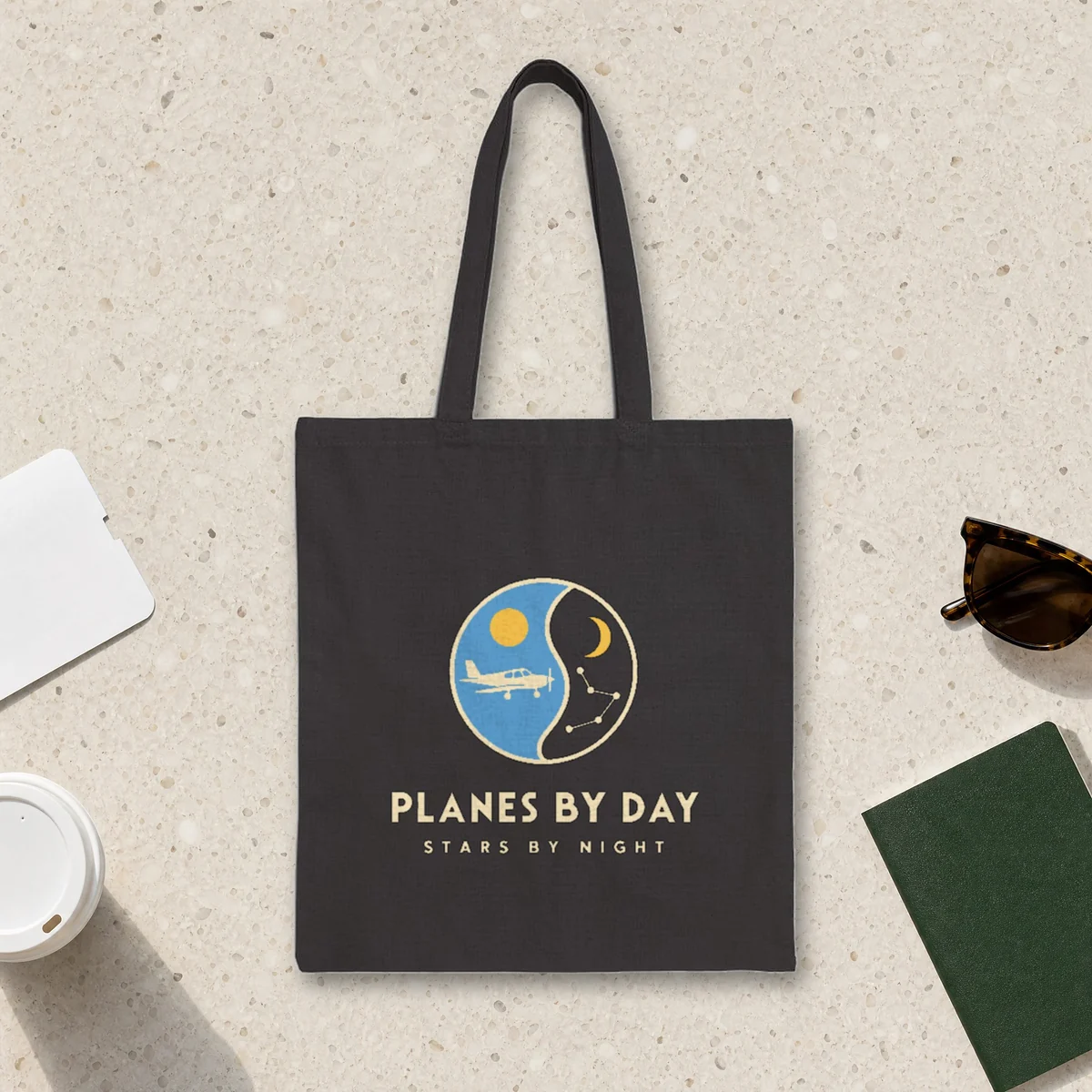 Planes by Day design in warm cream ink on a black canvas tote, laid flat on a light terrazzo terminal floor