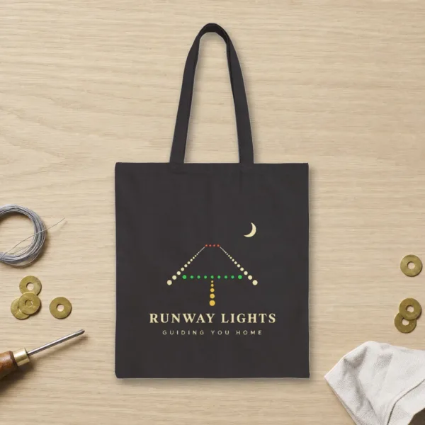 Runway Lights design in warm cream ink on a black canvas tote, laid flat on an oak hangar workbench