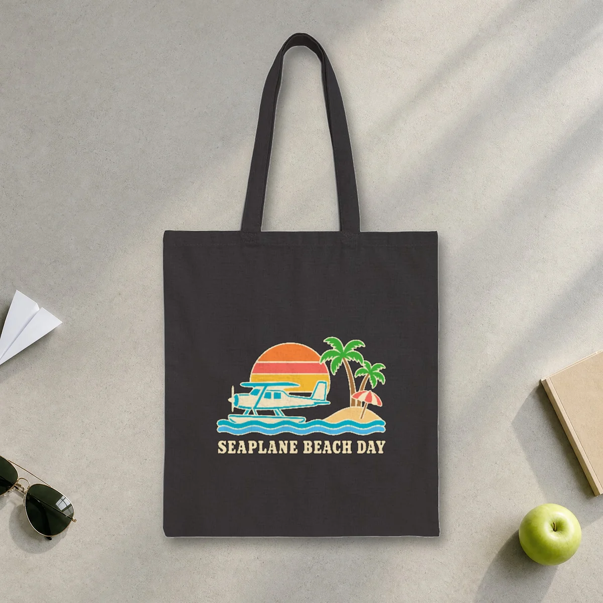 Seaplane Beach Day design in warm cream ink on a black canvas tote, laid flat on a sunlit terminal floor by the window