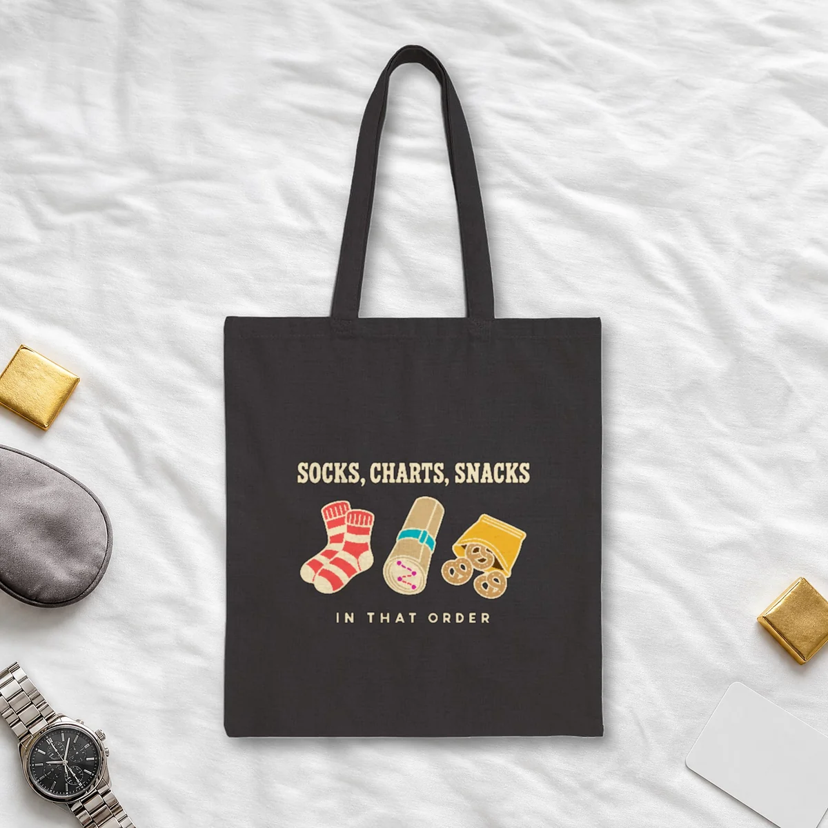 Socks Charts Snacks design in warm cream ink on a black canvas tote, laid flat on a crew hotel bedspread