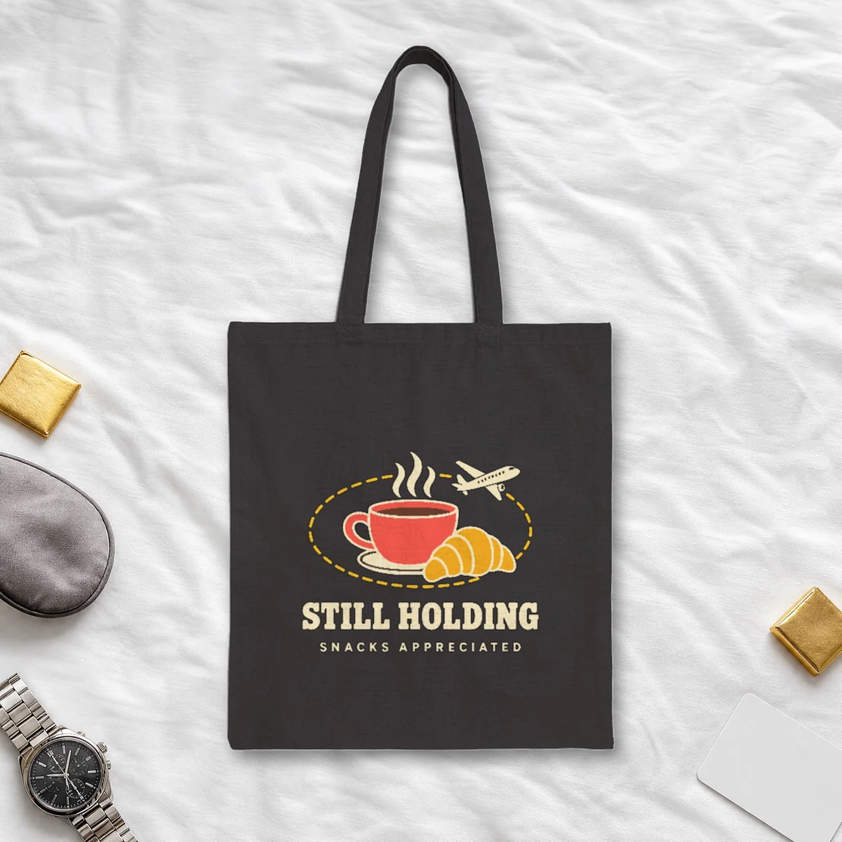 Still Holding design in warm cream ink on a black canvas tote, laid flat on a crew hotel bedspread