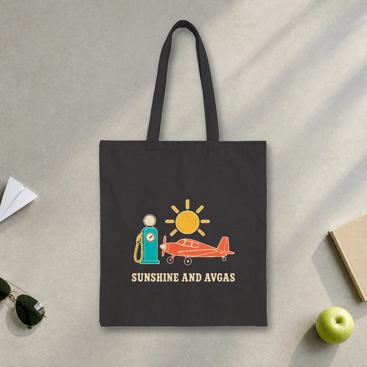 Sunshine and Avgas design in warm cream ink on a black canvas tote, laid flat on a sunlit terminal floor by the window