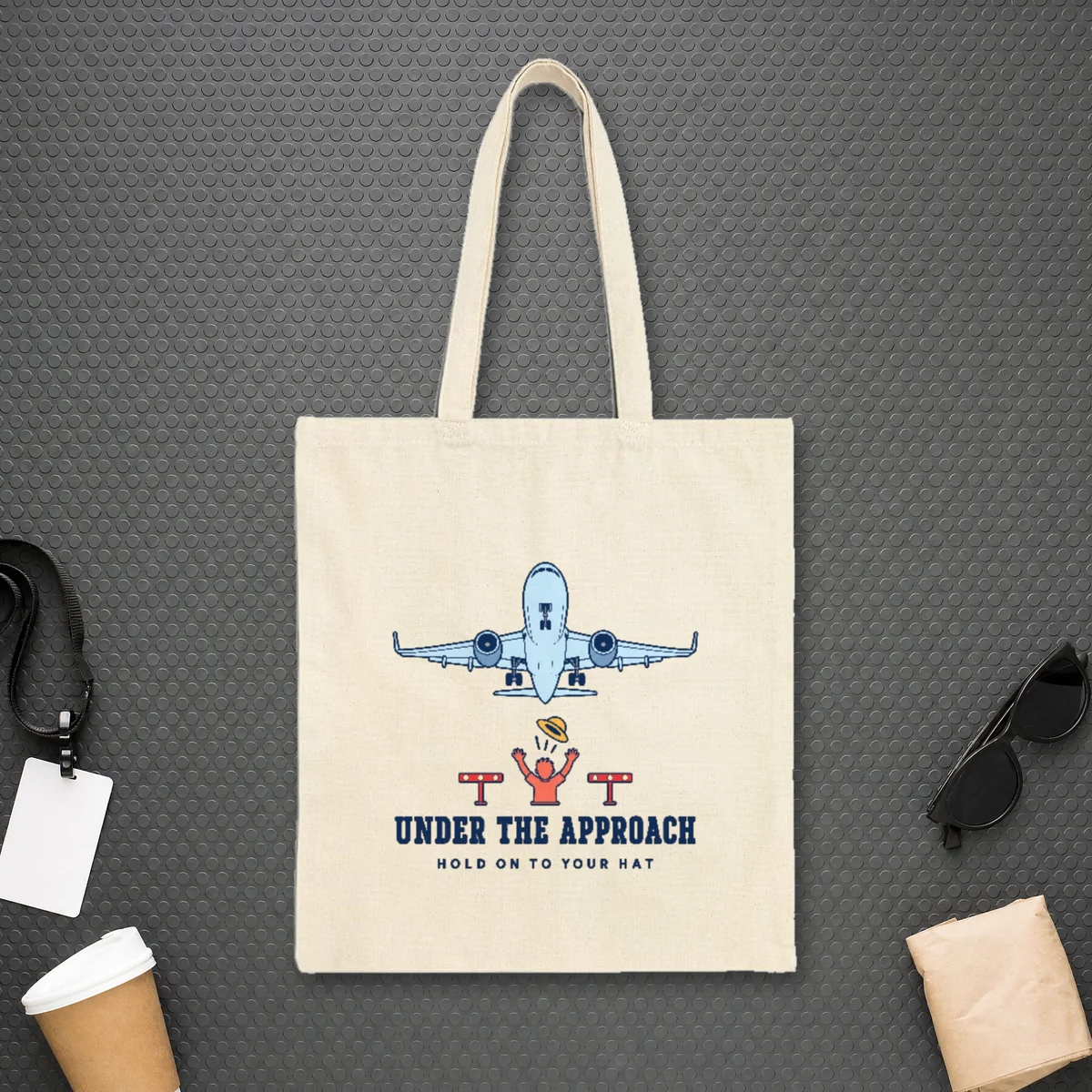 Under the Approach design in navy ink on a natural canvas tote, laid flat on the studded rubber floor of a jet bridge